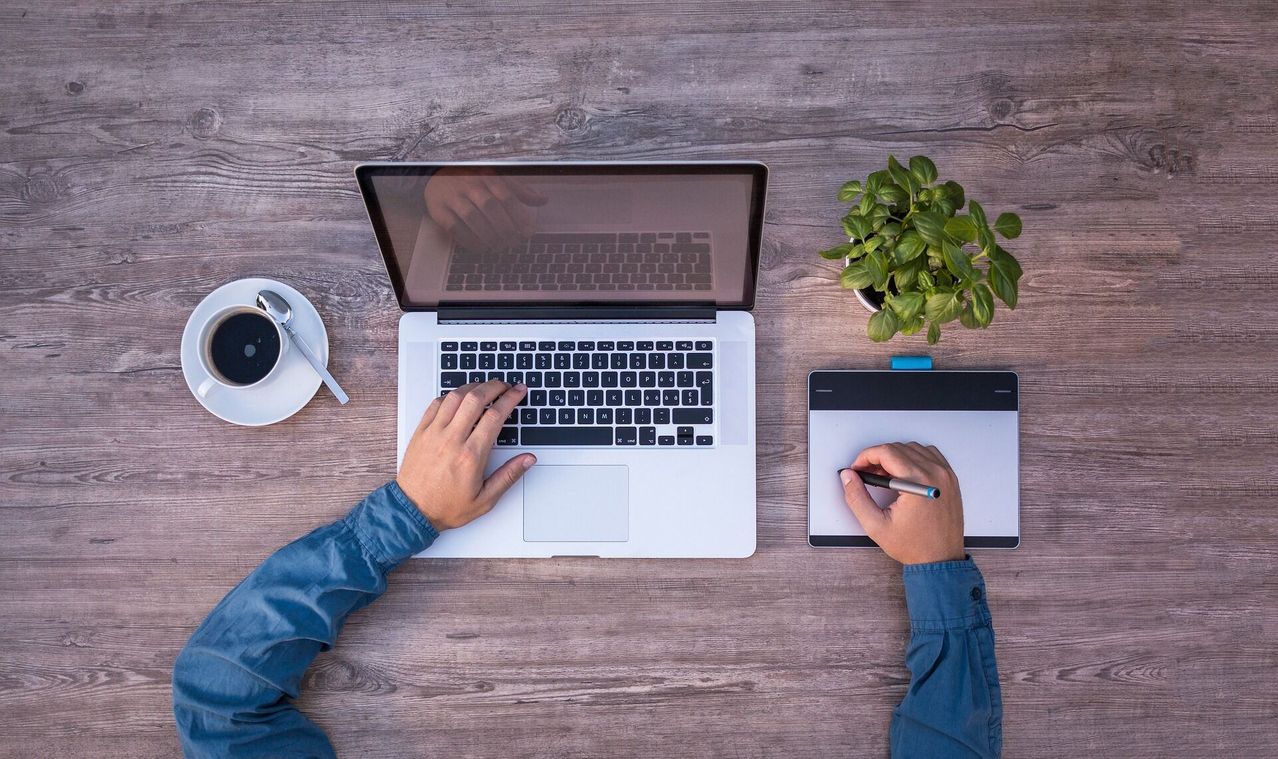 Eine Person arbeitet an einem Laptop, tippt auf der Tastatur und zeichnet auf einem Tablet. Eine Kaffeetasse steht auf dem Schreibtisch.