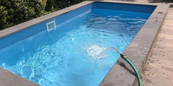 Ein leerer blauer Swimmingpool mit einem Abfluss am Boden. Ein Schlauch sprüht Wasser hinein. Ein Holzdeck umgibt ihn.