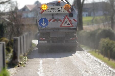 Ein Fahrzeug mit der Aufschrift '5m Abstand halten' fährt auf einer Straße mit Gras und Bäumen auf beiden Seiten.
