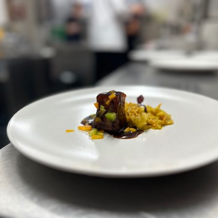 Ein Gourmetgericht auf einem weißen Teller, garniert mit dunkler Sauce, Gemüse und einem Hauptprotein. Der Hintergrund ist unscharf und zeigt eine Küche.