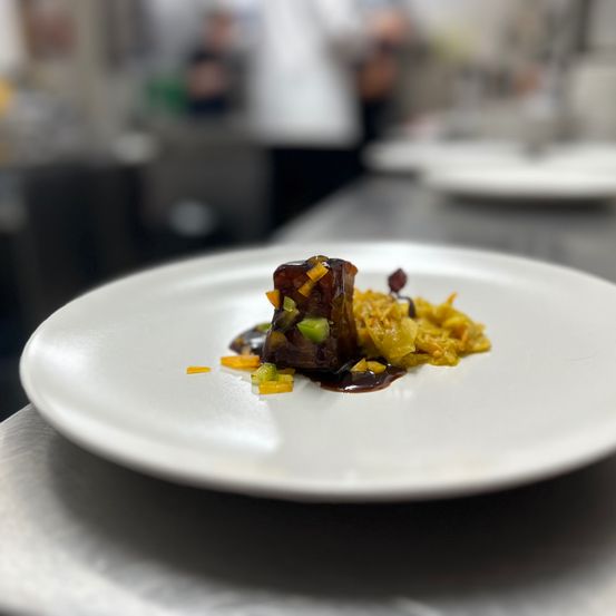 Ein Gourmetgericht auf einem weißen Teller, garniert mit dunkler Sauce, Gemüse und einem Hauptprotein. Der Hintergrund ist unscharf und zeigt eine Küche.