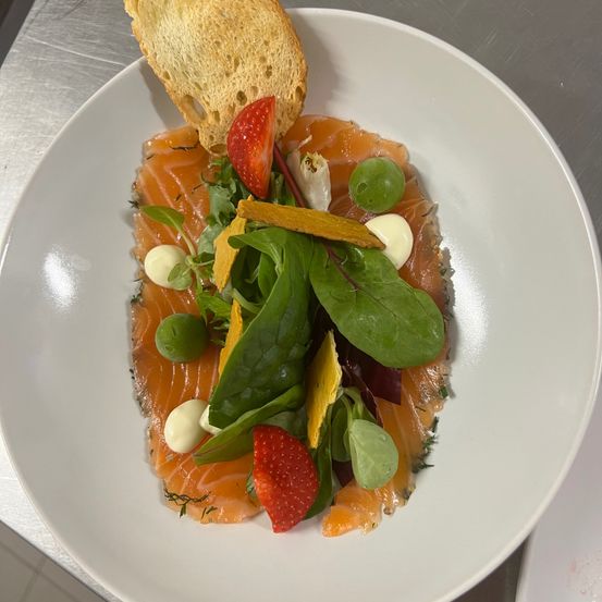 Ein weißer Teller zeigt einen Lachssalat mit Salat, Erdbeeren und einer Scheibe Brot.