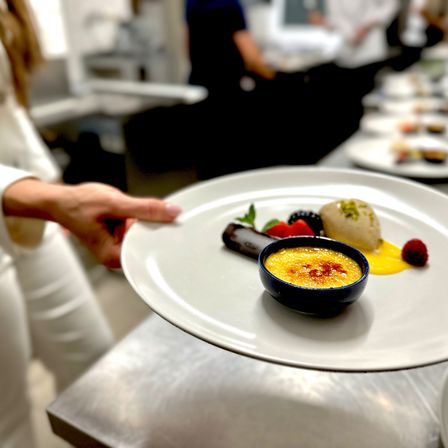 Eine Person in der Küche hält einen Teller mit einem Dessert. Das Dessert besteht aus einer Schüssel mit Crème brûlée, Beeren und einer Kugel Eis.