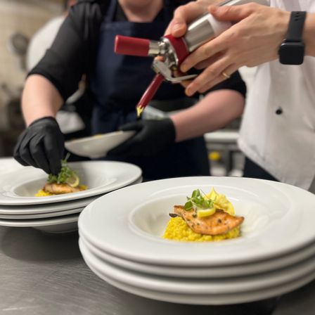 Zwei Köche in einer Küche, die Lachs mit Zitrone und Garnierung auf einem weißen Teller anrichten.