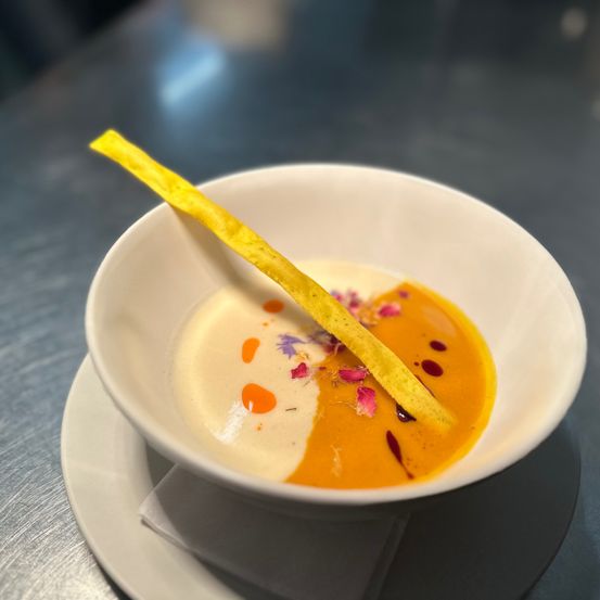 Ein weißer Teller auf einer Untertasse mit einer cremigen Suppe in orangefarbenem Sud, garniert mit Blütenblättern und einem gelben Stiel.