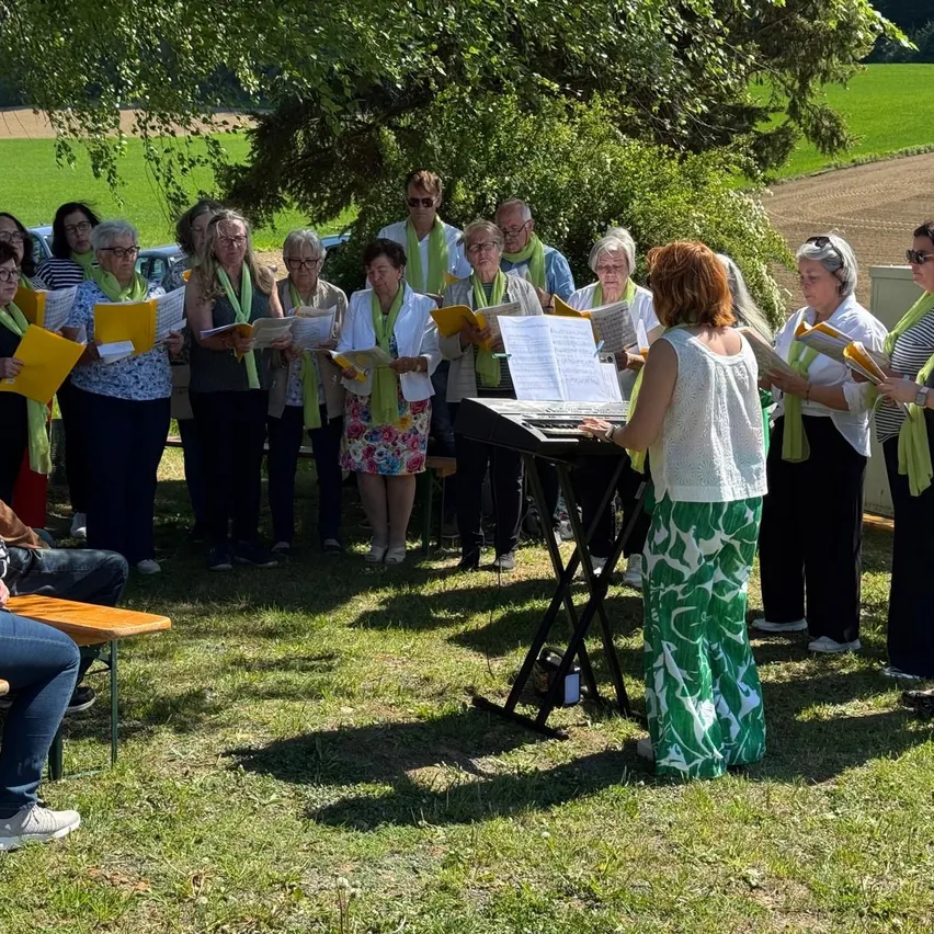 Ein Chor, gekleidet in grünen Schals, tritt im Freien unter einem Baum auf. Sie stehen vor einem Klavier mit Notenblättern. Hinter ihnen ist ein Feld und Autos sichtbar.