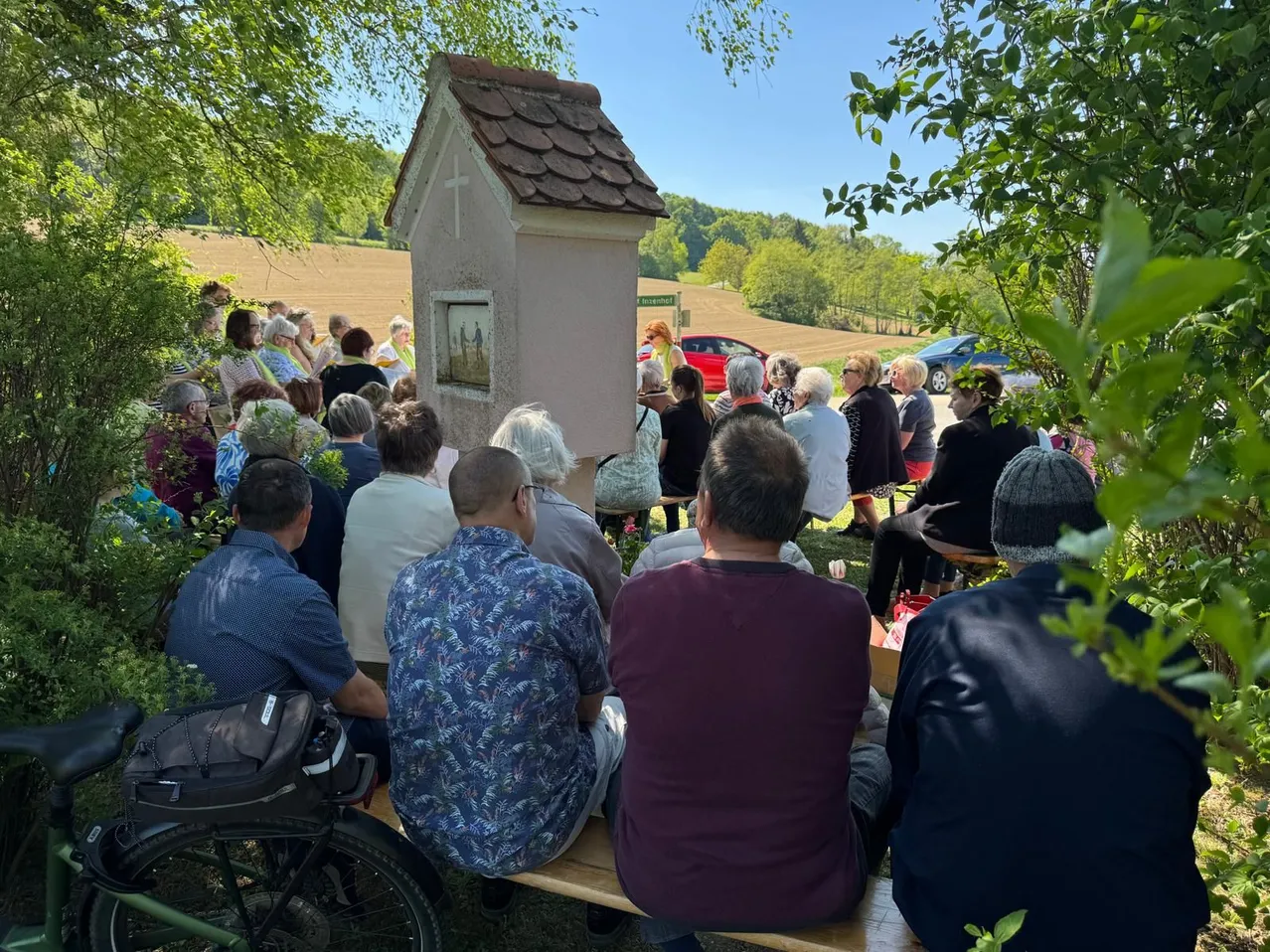 Eine Gruppe von Menschen sitzt auf Bänken unter Bäumen, mit einer Kapelle in der Mitte. Eine Person hält ein Kreuz. Ein rotes Auto und ein blaues Auto sind in der Nähe geparkt.