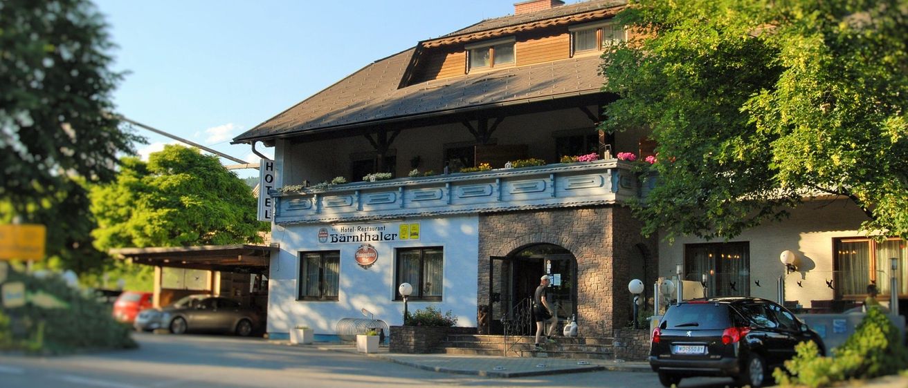 Ein Gebäude mit dem Schild Hotel Barnthaler, Blumen auf dem Balkon, eine eintretende Person. Ein Auto geparkt neben einem Baum, ein weiteres Auto auf der Straße geparkt.