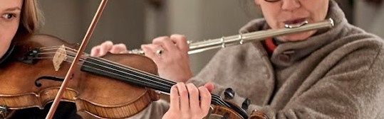 Zwei Frauen spielen Musikinstrumente, eine spielt Geige und die andere Flöte. Sie sind beide warm gekleidet in Winterkleidung.