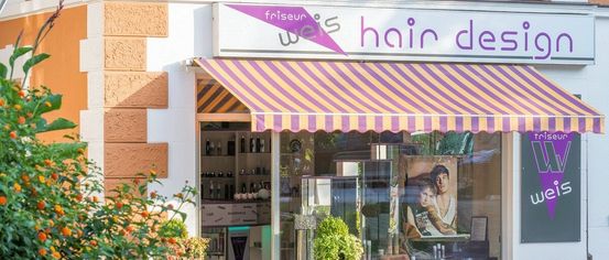 A store with a striped awning on a street. It has a sign reading 'friseur weis hair des'. The store has a glass door and window with a poster of a man and a woman.