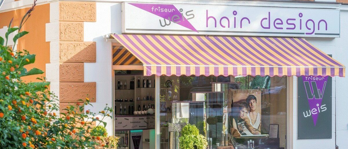 Ein Geschäft mit einem gestreiften Vordach an einer Straße. Es hat ein Schild mit der Aufschrift 'friseur weis hair des'. Das Geschäft hat eine Glastür und ein Fenster mit einem Poster eines Mannes und einer Frau.