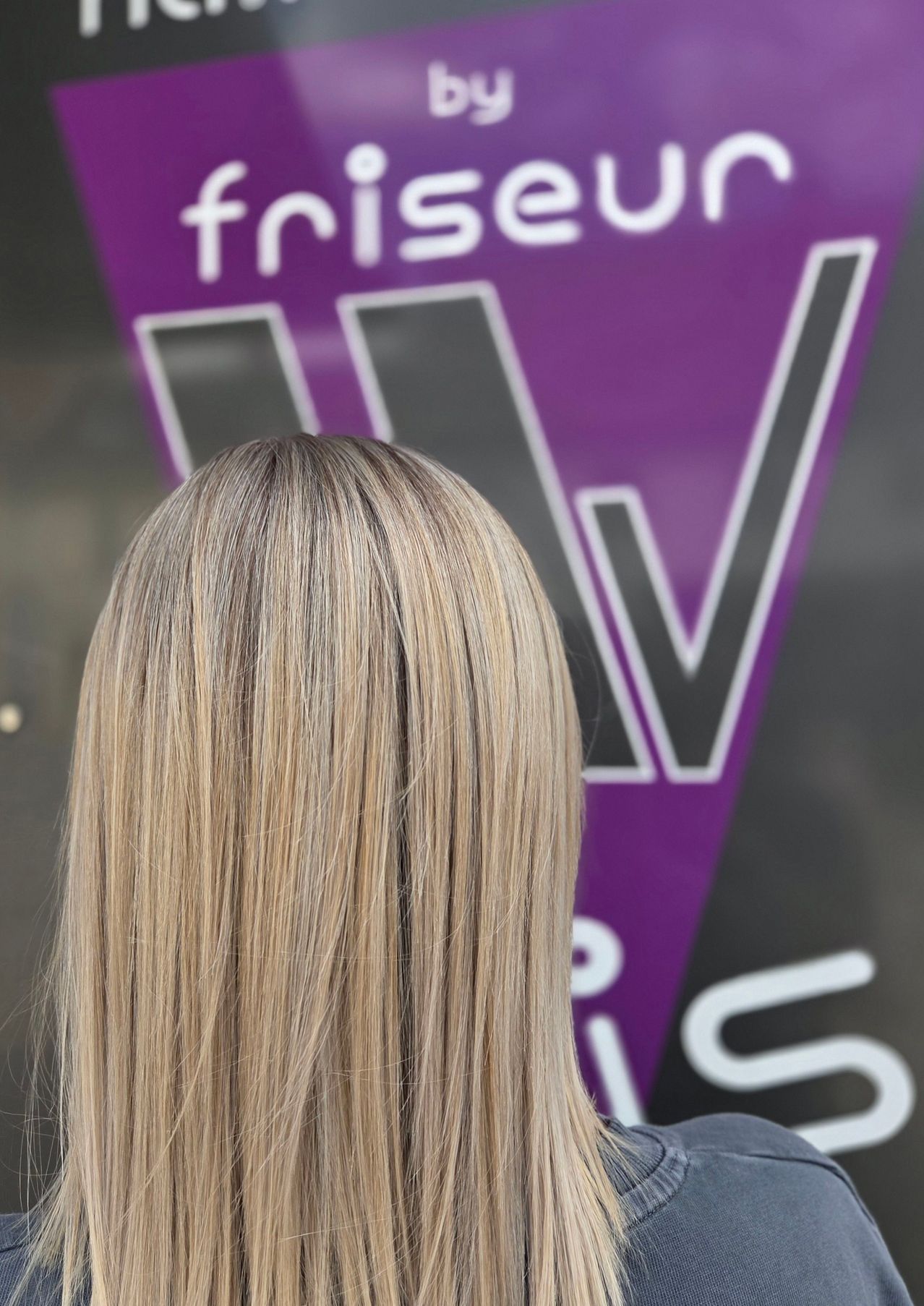 Eine Frau mit langen, hellblonden Haaren steht vor einem lila Banner mit den Buchstaben 'V' und 'W'.
