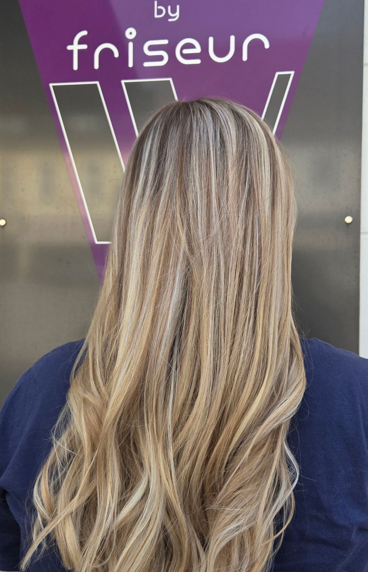 A woman with long, wavy blonde hair styled in a loose, natural look. She is wearing a blue shirt.