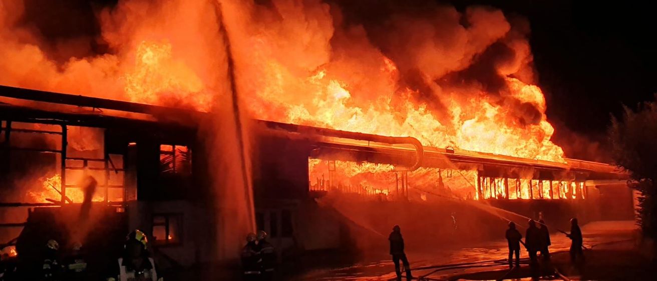 Feuerwehrleute kämpfen gegen ein massives Feuer in einem Gebäude. Flammen greifen das Bauwerk an und Rauch steigt in die Nacht auf.