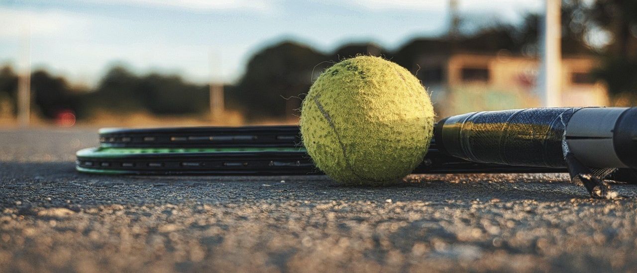 Ein Tennisball liegt auf einem Tennisplatz neben einem Schläger, unter einem teilweise bewölkten Himmel.