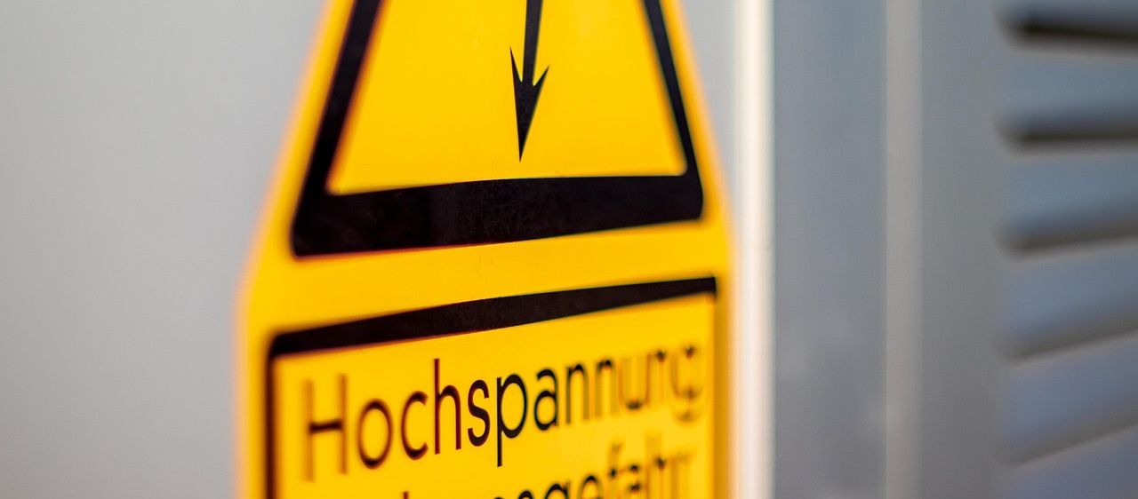 Ein gelbes Schild mit einem schwarzen Pfeil nach unten und den Worten Hochspannung Lebensgefahr.