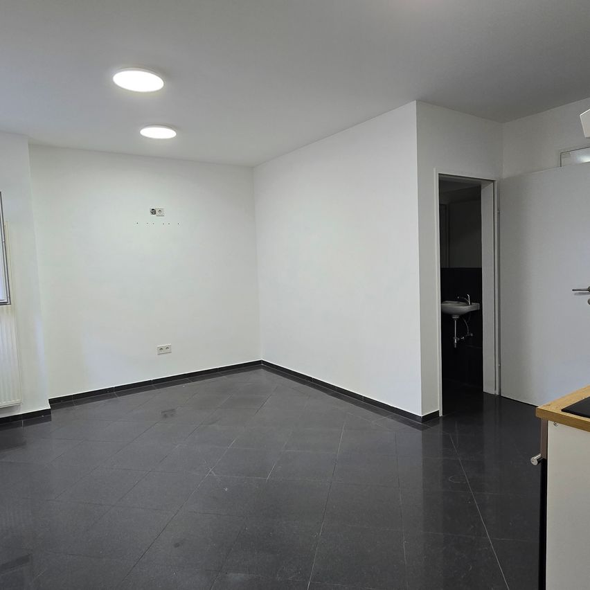An empty room with white walls, black tile floor, and a white door leading to a sink. There are two ceiling lights and a radiator on the left wall.