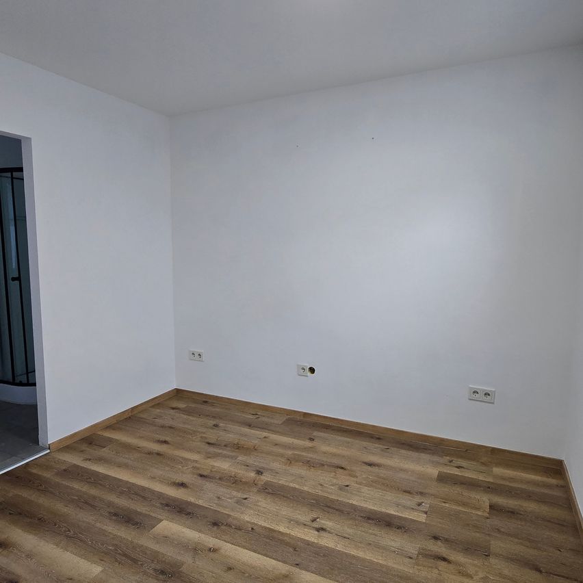 An empty room with white walls and wooden floor. There are electrical outlets on the walls and a door leading to another room.