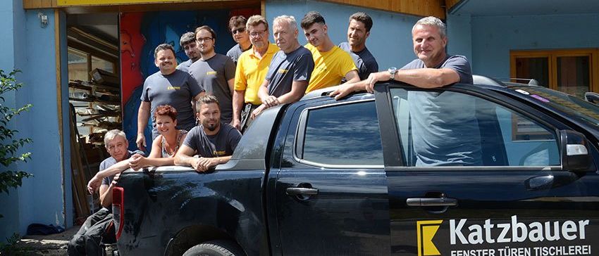 Eine Gruppe von Menschen posiert für ein Foto vor einem Gebäude mit einem gelben Schild, auf dem Katzbauer Fenster Türen Tischlerei steht. Sie stehen auf einem schwarzen Pickup-Truck.