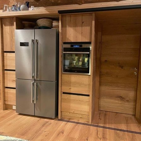 Eine moderne Küche mit Holzschränken, einem Edelstahlkühlschrank und einem in die Schränke integrierten Ofen. Der Raum verfügt über Holzböden und eine rustikale Wand.