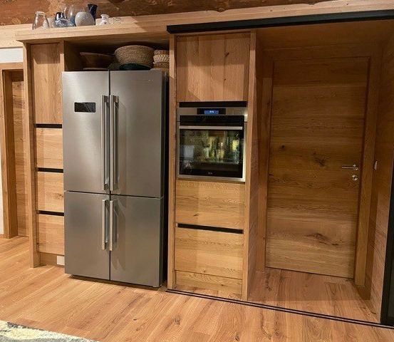 Eine moderne Küche mit Holzschränken, einem Edelstahlkühlschrank und einem in die Schränke integrierten Ofen. Der Raum verfügt über Holzböden und eine rustikale Wand.