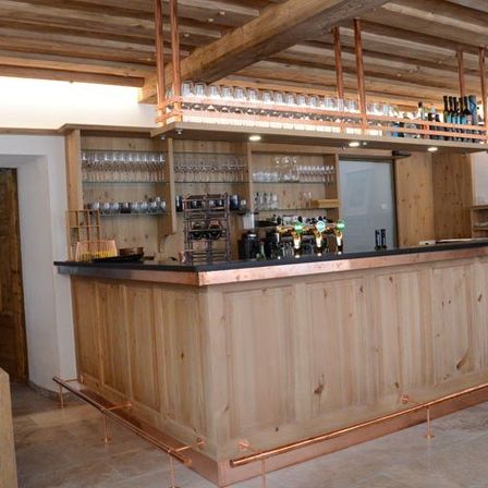 Eine Holztheke mit Kupferakzenten und Regalen darüber, mit Weinläsern, Flaschen und einem Zapfhahn. Die Bar befindet sich in einem rustikalen Raum mit Holzbalken und gefliesten Böden.