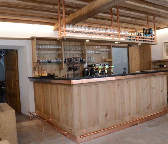 Eine Holztheke mit Kupferakzenten und Regalen darüber, mit Weinläsern, Flaschen und einem Zapfhahn. Die Bar befindet sich in einem rustikalen Raum mit Holzbalken und gefliesten Böden.