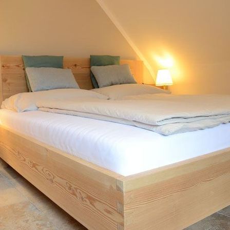 Ein ordentlich gemachtes Bett mit weißer Bettwäsche und Kissen, Holzbettrahmen und zwei Lampen auf beiden Seiten. Ein Holzstuhl ist neben dem Bett platziert.