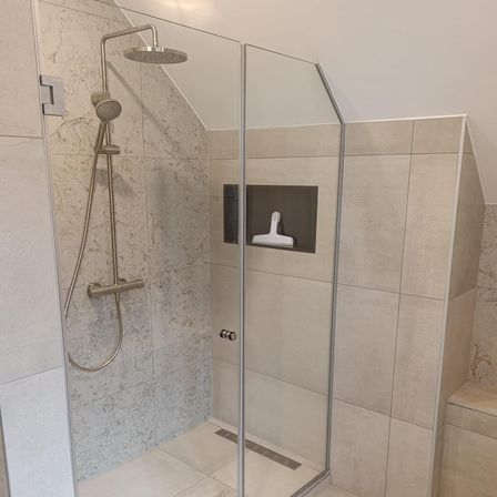 Ein moderner Duschbereich mit einer Glastür, gefliesten Wänden und einer eingebauten Duschbrause mit Schlauch. Die Duschbrause ist silber und die Wände sind beige mit Marmormuster.