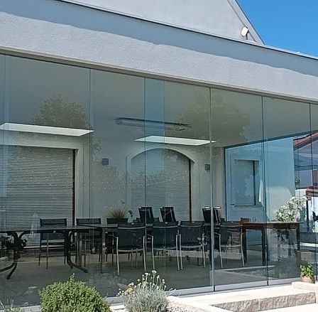 Ein modernes Haus mit Glaswänden und -türen zeigt einen Innenbereich mit einem langen Tisch und Stühlen. Blumentöpfe und Blumen sind draußen.