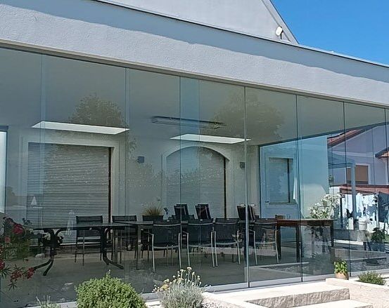 Ein modernes Haus mit Glaswänden und -türen zeigt einen Innenbereich mit einem langen Tisch und Stühlen. Blumentöpfe und Blumen sind draußen.