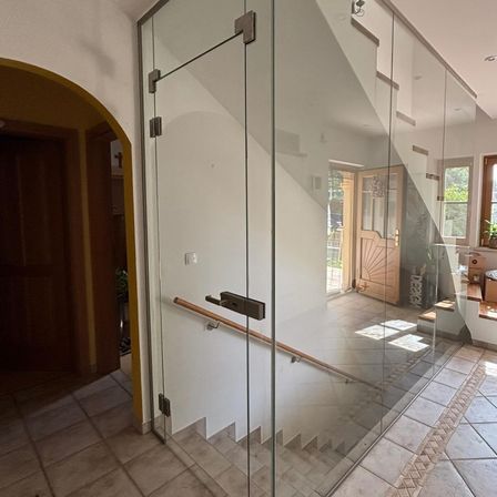 Eine moderne Treppe mit Glaspaneelen und einem Holzhandlauf. Die Treppe führt zu einer Holztür mit einem Glasfenster und einem Topf mit Pflanzen.