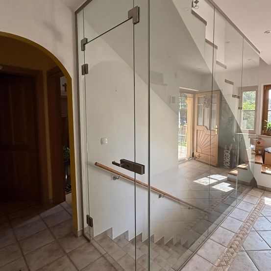 Eine moderne Treppe mit Glaspaneelen und einem Holzhandlauf. Die Treppe führt zu einer Holztür mit einem Glasfenster und einem Topf mit Pflanzen.