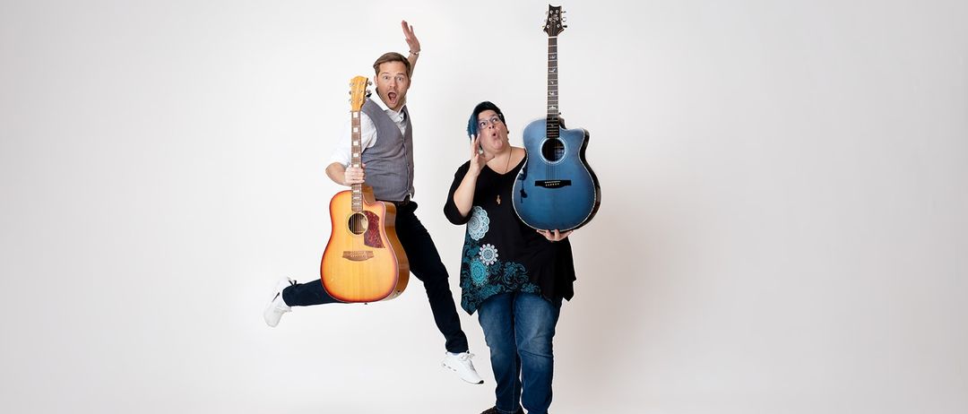 Ein Mann und eine Frau springen zusammen und halten Gitarren. Der Mann ist in der Luft, während die Frau auf einer Holzplanke steht. Beide lächeln.