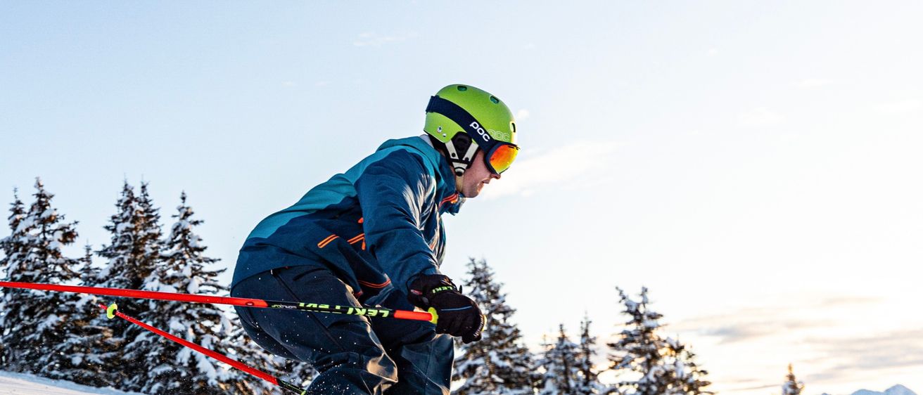 Ein Skifahrer mit Schutzbrille und grünem Helm fährt einen schneebedeckten Hang hinunter, im Hintergrund stehen Tannen.