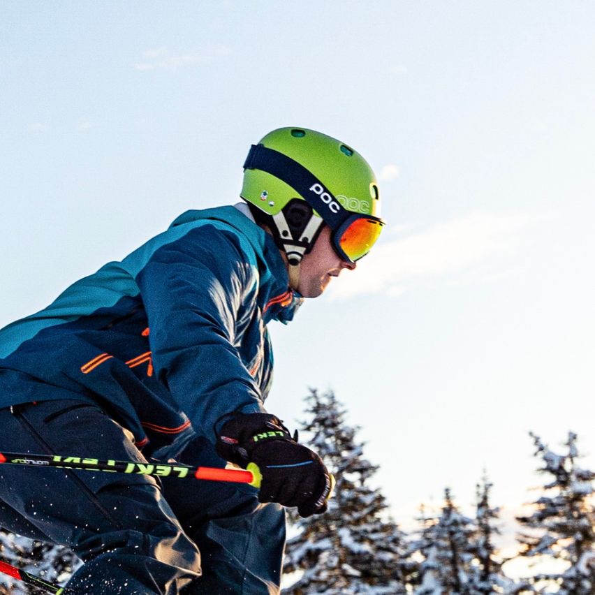 Ein Skifahrer mit Schutzbrille und grünem Helm fährt einen schneebedeckten Hang hinunter, im Hintergrund stehen Tannen.