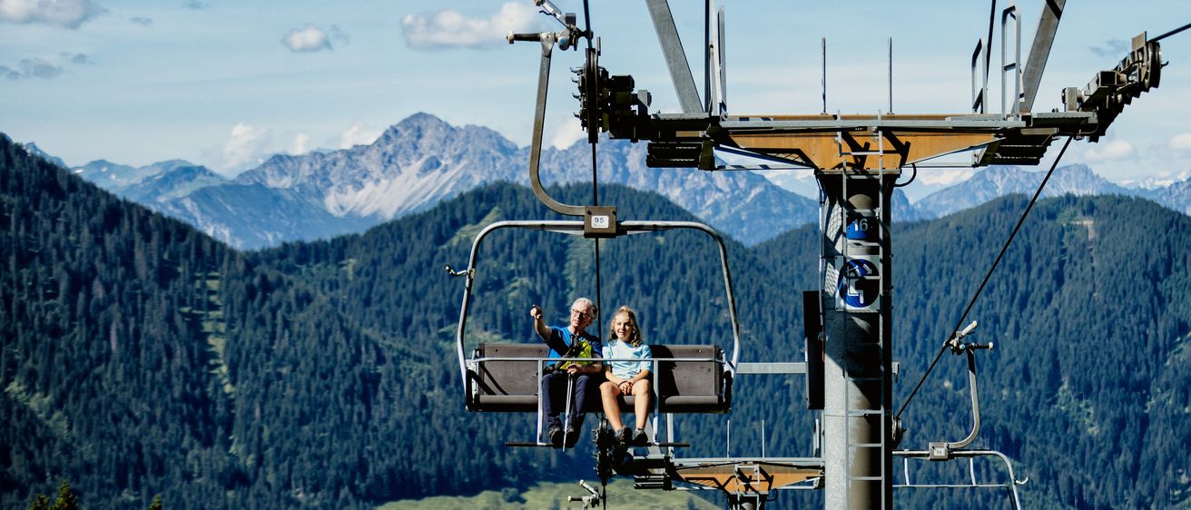 Ein älteres Paar fährt mit Blick auf die Alpen mit einer Skilift. Unten trägt ein weiterer Lift drei Personen.