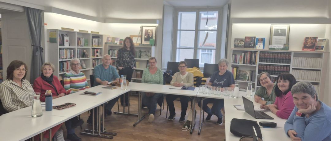 Gruppe von Menschen, die in einem Konferenzraum an Tischen sitzen, mit Bücherregalen im Hintergrund.
