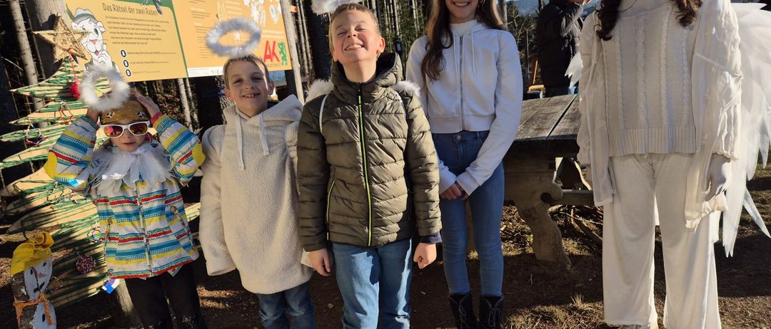 Eine Gruppe von Kindern mit Engelflügeln und Winterkleidung stehen draußen. Ein Schild steht hinter ihnen, und ein Mann steht in der Nähe einer Bank.