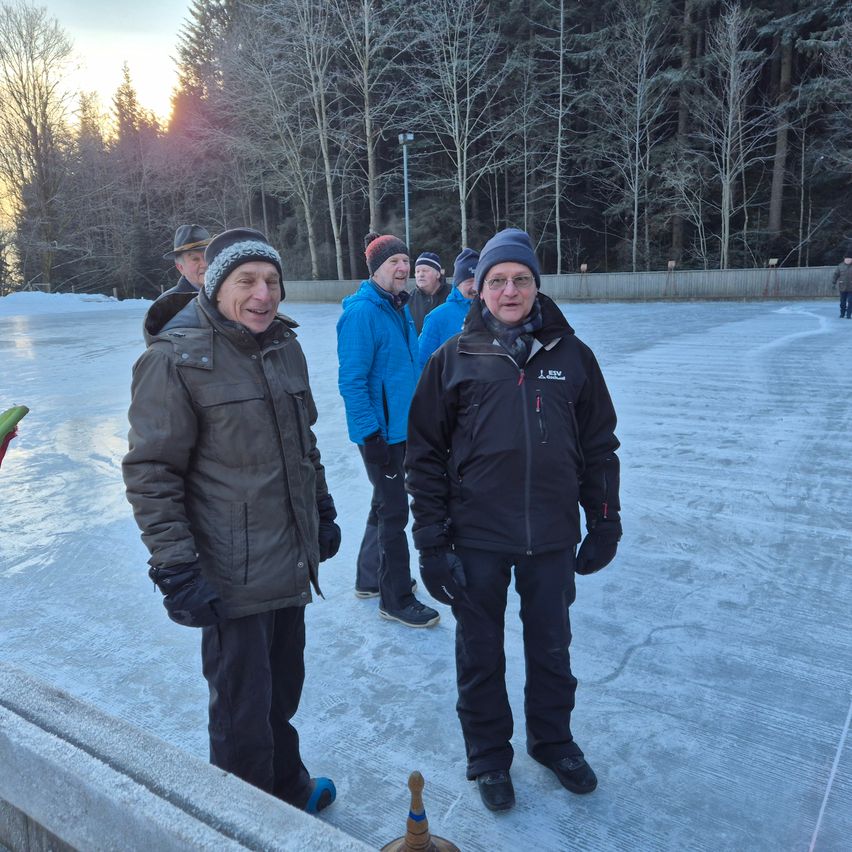A group of people bundled up in winter clothing stand on an outdoor ice rink, with a man in glasses smiling and another in a black jacket facing the camera.