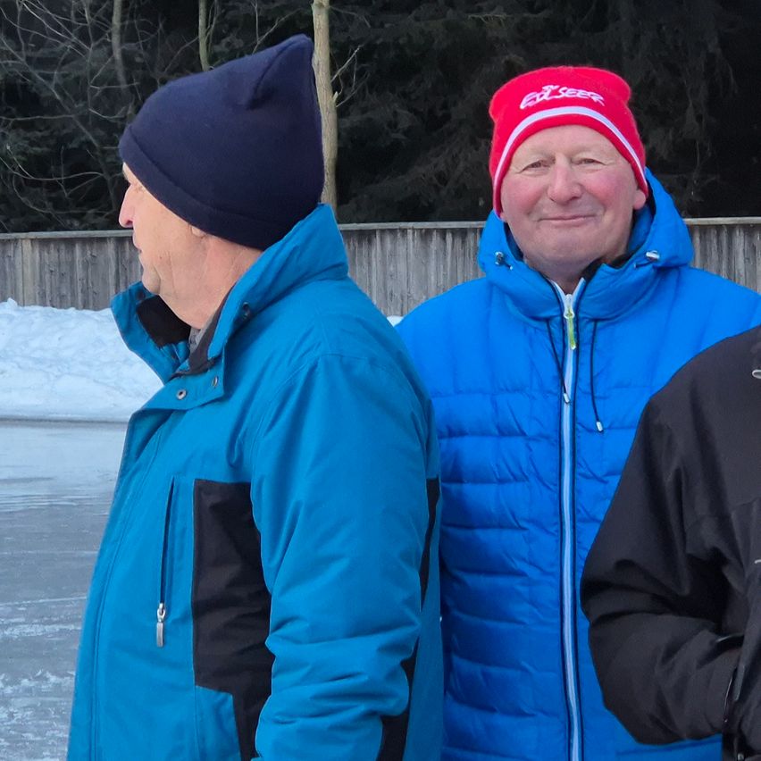 Two men in blue jackets and beanies stand near a snowy fence, smiling at the camera.