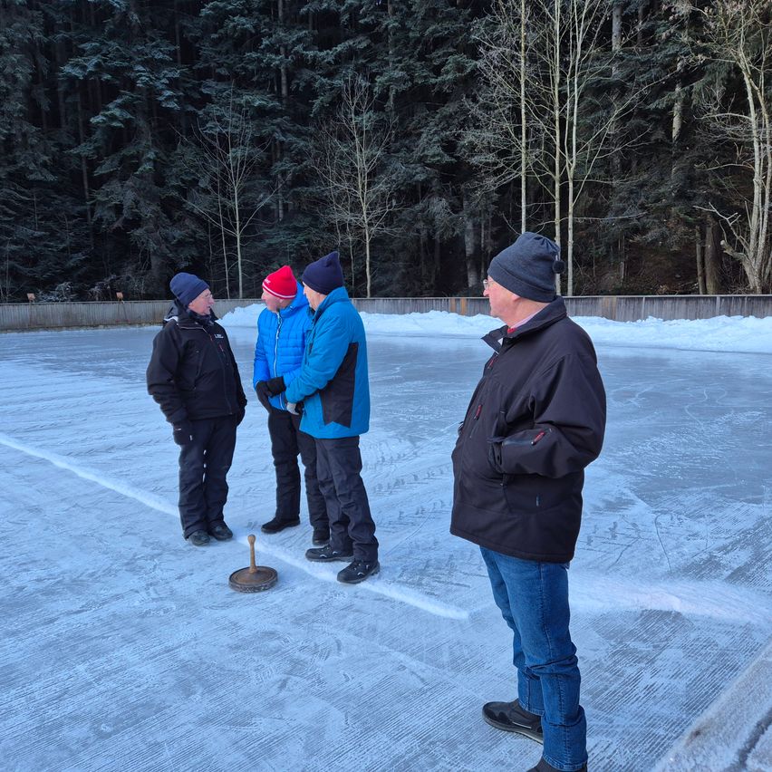 Four men stand on an ice rink, discussing something. They are all wearing winter clothes and hats. A puck is on the ice.