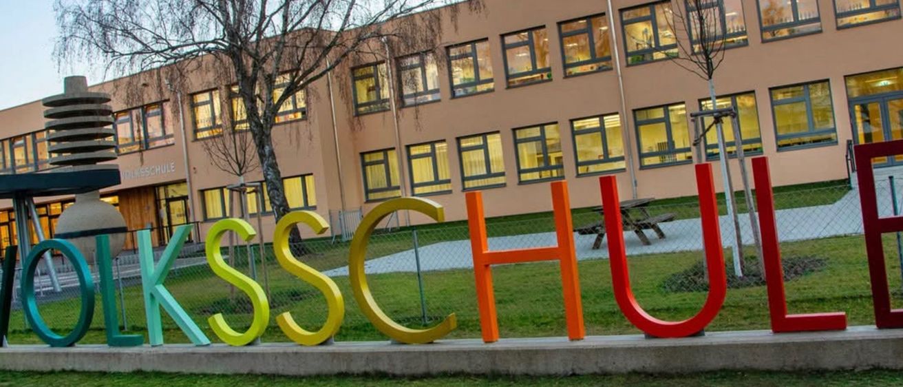 Ein Gebäude mit vielen Fenstern, das große Buchstaben S, C, H, U auf dem Rasen davor aufweist. Ein Baum und eine Bank im Hintergrund.