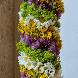Eine hohe Blumenarrangement mit weißen, gelben und lila Blumen, einschließlich Gänseblümchen, vor einem strukturierten Wandhintergrund.