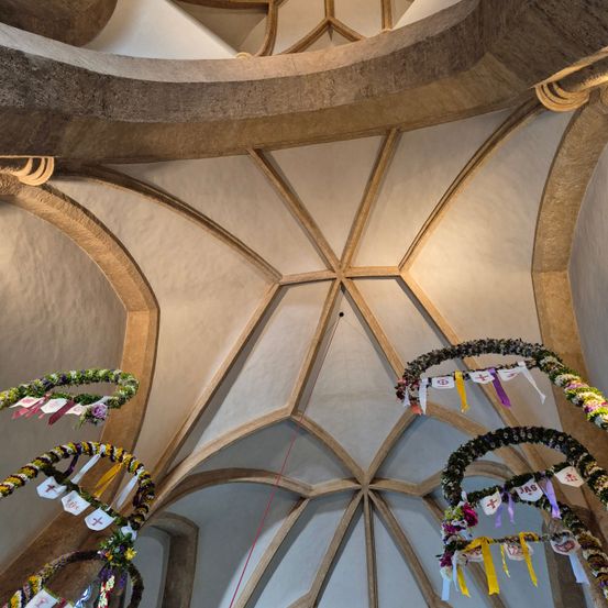 Das Innere einer Kathedrale mit dekorativen Blumenkränzen, die von der Decke hängen. Die Decke hat ein sternförmiges Design mit geometrischen Mustern. Die Wände sind gewölbt mit Bögen.