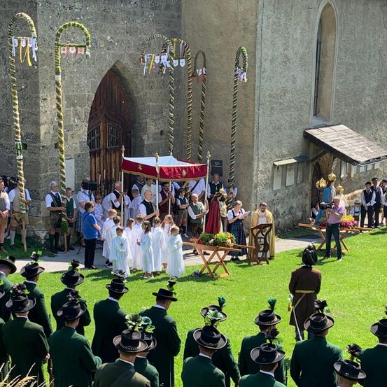 Eine religiöse Zeremonie vor einer Kirche mit einem Baldachin und einer Gruppe von Menschen in traditioneller Kleidung. Ein Mann steht an einem Podium, und andere beobachten.