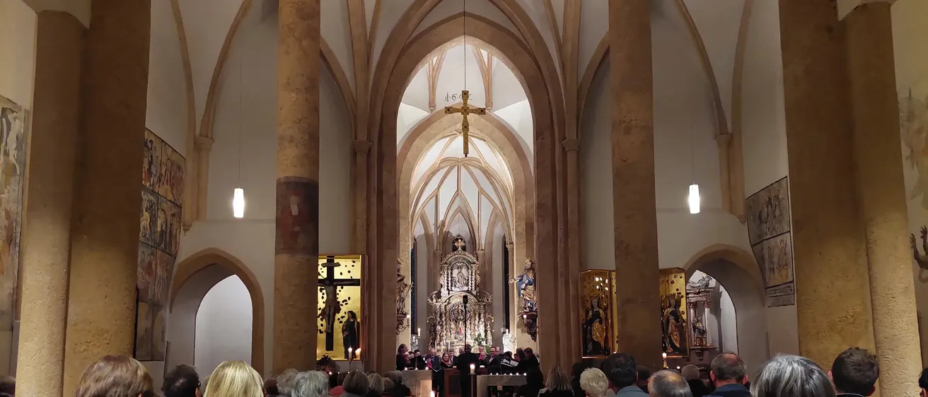 Eine Gruppe von Menschen nimmt an einem Gottesdienst in einer Kirche teil. Sie sitzen auf hölzernen Kirchenbänken, die zum Altar ausgerichtet sind, wo ein Chor singt. Der Altar hat ein Kreuz, Kerzen und religiöse Kunstwerke.