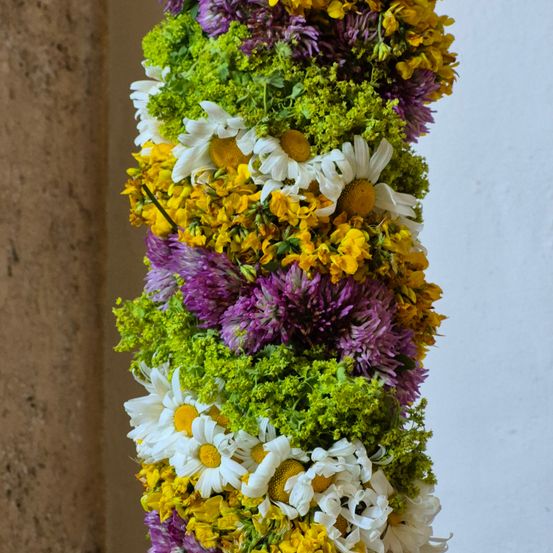 Eine vertikale Blumenanordnung mit einer Mischung aus weißen, gelben und lila Blumen. Die Blumen sind in einem kreisförmigen Muster mit grünen Blättern angeordnet. Die Blumen werden wahrscheinlich zur Dekoration verwendet.
