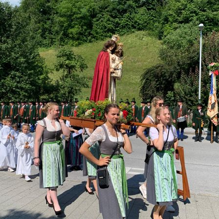 Mehrere Frauen, die eine Statue einer religiösen Figur tragen, in traditionellen grünen Kleidern gekleidet, gehen an einer Menschenmenge in grünen Uniformen an einem sonnigen Tag vorbei.