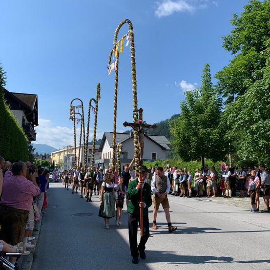 Ein Straßenumzug mit Menschen in traditioneller bayerischer Kleidung, die große, mit Blumen dekorierte Stangen und ein Kreuz tragen, unter einem blauen Himmel mit Bergen in der Ferne.
